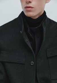 Giacca in misto lana nera con colletto alto, chiusura frontale con bottoni e due tasche sul petto; indossata sopra un maglione a collo alto nero.