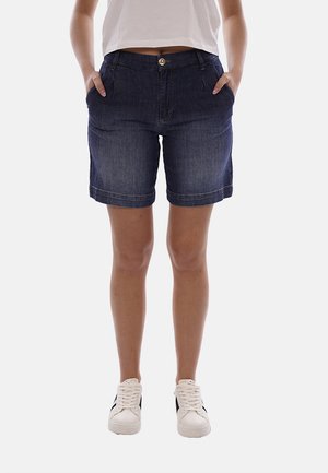 Persona che indossa pantaloncini di denim blu scuro, sneakers bianche e una maglietta bianca, in piedi con le mani in tasca contro uno sfondo semplice.