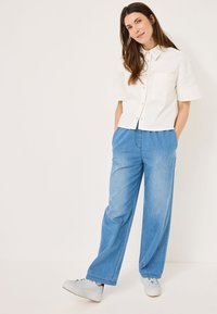 Camicia bianca a maniche corte con bottoni e taschino, abbinata a pantaloni di denim azzurri a gamba larga e sneakers grigie con dettagli rossi.