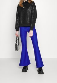 Sudadera negra con estampado y capucha con cordón, combinada con pantalones azules de textura y acampanados. Los accesorios incluyen un pequeño bolso de mano plateado.
