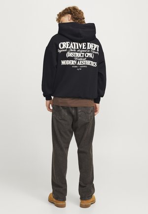 Black hooded sweatshirt featuring large white text on the back. Paired with brown long-sleeve shirt and dark grey relaxed-fit jeans. Brown boots.