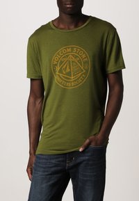 Grøn bomulds t-shirt med et cirkulært grafisk motiv med teksten "VOLCOM STONE DRIFTERS SOCIETY" i gul. Korte ærmer og rund halsudskæring.