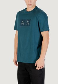 T-shirt en coton teal avec un graphique rectangulaire de couleurs sombres comportant le texte "AX" et "ARMANI EXCHANGE". Col rond côtelé et manches courtes.