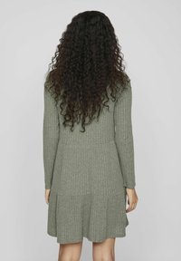 Personne aux longs cheveux bouclés foncés portant une robe grise côtelée à manches longues, arrivant aux genoux, vue de dos sur un fond uni.