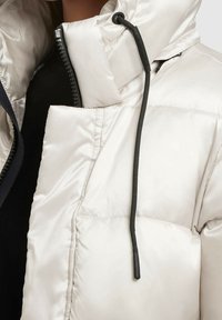 Chaqueta acolchada de color claro con cuello alto, cremallera negra, detalles de cordón y textura acolchada. Material suave y brillante.