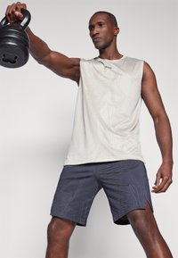 Camisa sin mangas de color beige claro con textura estampada y logo, combinada con shorts oscuros de color navy con diseños geométricos y aberturas laterales.