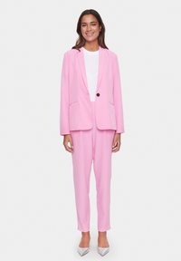 Růžový přizpůsobený blazer s výstřihem do V a jedním knoflíkem, kombinovaný s odpovídajícími zúženými kalhotami a bílým topem, doplněný stříbrnými podpatky.