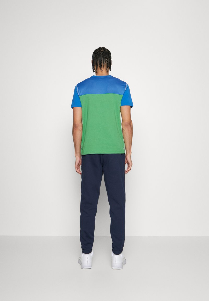 T-shirt à manches courtes avec un corps vert, des épaules bleues et une finition texturée, associé à un jogging navy et des baskets blanches.