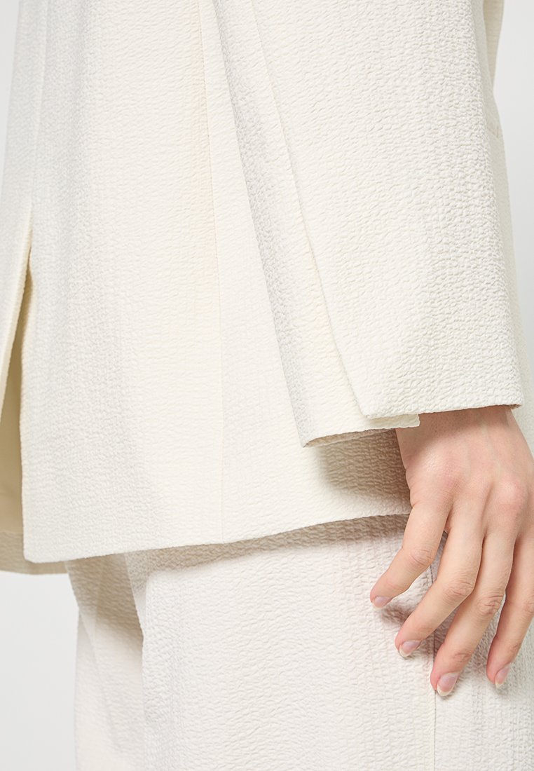 Weißer strukturierten Blazer mit leicht übergroßer Passform und strukturiertem Design. Der Stoff hat eine zerknitterte Oberfläche und wird mit passenden Hosen kombiniert.