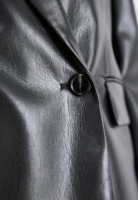 Giacca in pelle nera con superficie texture, dotata di chiusura a un bottone e rever su misura. Linee nette e finitura liscia.