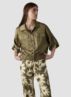 Frau trägt olivgrüne Knopfleiste Jacke mit hochgekrempelten Ärmeln und elastischem Bund, kombiniert mit grün-weißen Blumenmuster weiten Hosen.