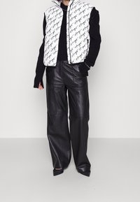 Bílá vatovaná vesta s černým logem, doplněná černými širokými koženými kalhotami. Pod ní je černý texturovaný pletený svetr.