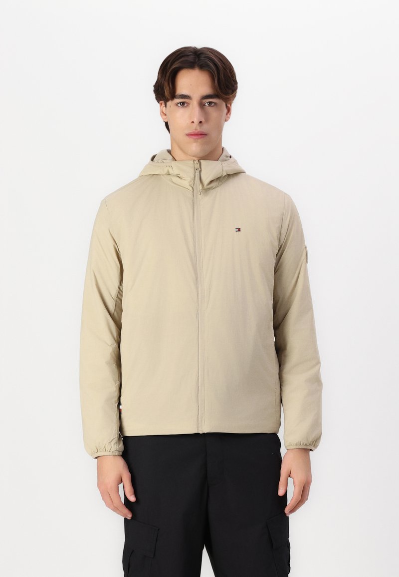 Lys beige zip-jakke med hette, elastiske mansjetter og en liten logo på brystet. Brukt over svarte cargo-bukser.