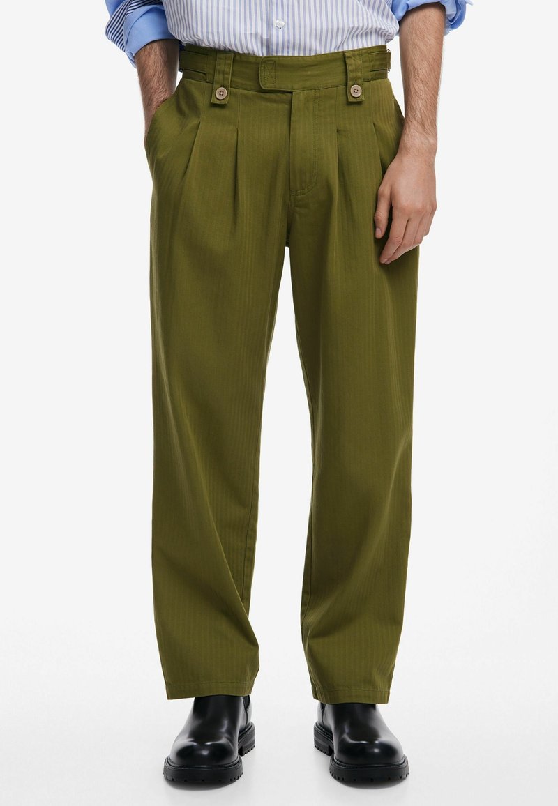 Homme portant un pantalon plissé vert olive avec des détails boutons et des bottes en cuir noir, associé à une chemise rayée bleue à manches retroussées.