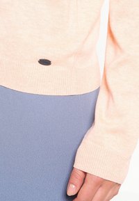 Pull en maille pêche clair avec une manche à bords-côtes et un petit logo noir en bas, associé à un bas texturé bleu clair.