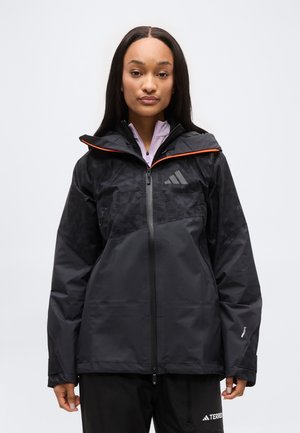 Vrouw die een zwarte en donkergrijze outdoorjas met rits, capuchon en oranje rand draagt, staand tegen een effen lichte achtergrond.