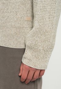 Μπεζ πουλόβερ με υφή, με ριμπ μανσέτες και τελείωμα, που διαθέτει μια μικρή δερμάτινη ετικέτα με λογότυπο. Φορεμένο πάνω από καφέ υφασμάτινο κάτω μέρος.