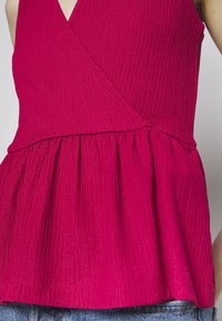 Femme portant un top cache-cœur sans manches texturé magenta avec une taille froncée, associé à un jean en denim bleu.