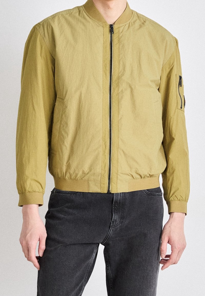 Homme portant une veste bomber marron clair avec une fermeture éclair devant et un jean noir, debout devant un fond clair uni.