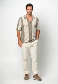 Camicia a maniche corte con bottoni a strisce beige e marrone, abbinata a pantaloni crema e scarpe stringate marroni. Il modello sta in piedi con sicurezza.