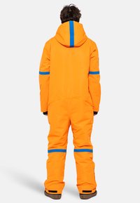 Tuta da sci arancione con motivo a strisce blu, materiale impermeabile e cappuccio. Include polsini elastici e cerniera alla caviglia.