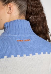 Blå og hvit strikket genser med høy ribbestrikket krage. Har et mønster nederst og "KARI TRAA" brodert i oransje på baksiden.