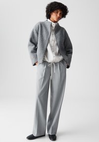 Giacca grigia di lana con texture a maglia, camicia bianca con bottoni e pantaloni a pieghe grigi chiari. Scarpe nere completano l'outfit.