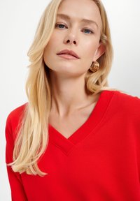Maglione rosso a scollo a V realizzato in tessuto a maglia, con dettagli a coste lungo il collo. Capelli lunghi e biondi acconciati in modo leggero vicino al collo.