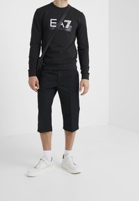 Felpa nera in cotone con un logo grafico a contrasto, abbinata a pantaloni corti neri fino al ginocchio e sneaker bianche con dettagli beige.