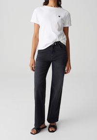 White cotton t-shirt with a small black heart logo, paired with high-waisted black jeans and black slide sandals.