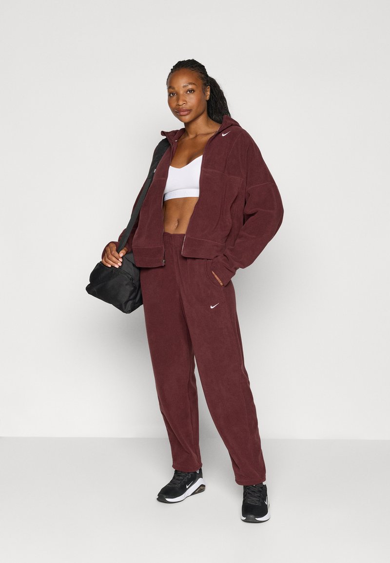 Tuta in pile bordeaux con giacca con cappuccio e pantaloni abbinati. Sotto, un reggiseno sportivo bianco, una borsa a tracolla nera e sneakers nere.