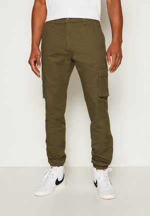 Homme portant un pantalon cargo vert olive avec poches à rabat sur les côtés et des baskets montantes blanches avec des logos swoosh noirs.