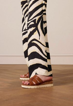 Brown leather sandals with two straps, light-colored wedge sole, paired with black and white striped wide-leg pants against wood flooring.