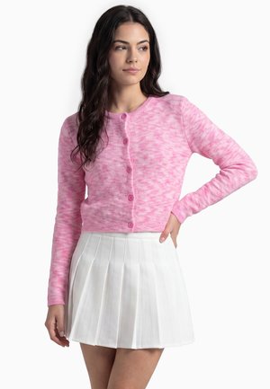 Donna con capelli lunghi e scuri che indossa un cardigan rosa con bottoni e una mini gonna bianca plissettata, posando con una mano sul fianco.