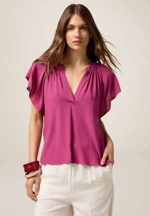 Fuchsia blouse met korte mouwen en een V-hals, voorzien van een geplooide halslijn, gemaakt van lichtgewicht stof, gecombineerd met witte, loose-fitting broeken.