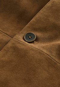 Brunt ruskind med et strømlinet design og en rund, mørk knap syet på en vandret linje, der forbedrer tekstur og detaljer.