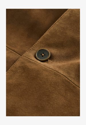 Braunes Wildledergewebe mit einem eleganten Design und einem runden, dunklen Knopf, der auf einer horizontalen Linie genäht ist und die Textur und Details hervorhebt.