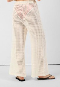 Crèmekleurige, opengehaakte brede broek met een textuurpatroon, die een contrasterende rode driehoek eronder onthult. Vlakke sandalen zichtbaar.