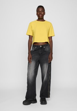 Gul croppet t-skjorte kombinert med løse svarte jeans som har en falmet design og en synlig glidelås. Svarte joggesko fullfører antrekket.