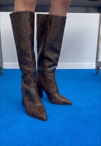 Nohy v hnědo-černých kožených vysokých elegantních botách s špičatou špičkou stojí na jasně modrém koberci.