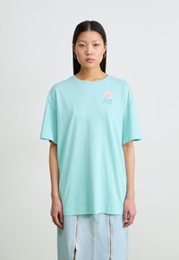 Tricou din bumbac verde mentă, cu mânecă scurtă, guler rotund și un design mic de logo roz pe partea stângă a pieptului.