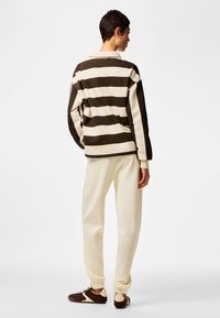 Oseba, oblečena v bež in temno rjavo črtasto dolgo majico, krem barve trenirke in temno rjave superge, stoji pred enobarvnim belim ozadjem.