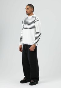 Wit overhemd met lange mouwen en horizontale zwarte strepen over de borst en armen, gecombineerd met losse zwarte broek en zwarte schoenen met accenten.