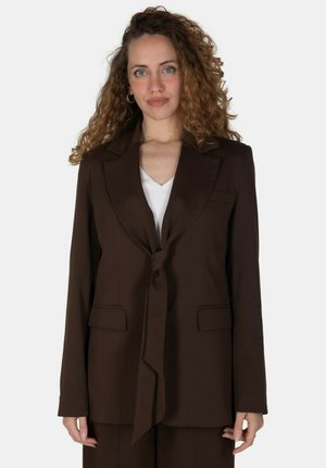 Donna con capelli ricci che indossa una giacca marrone annodata in vita sopra una maglietta bianca, in piedi davanti a uno sfondo semplice.