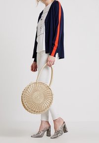 Kulatá, tkaná slaměná kabelka s kruhovým designem a texturovaným povrchem. Ruční nošení s pevným, přírodně zbarveným uchem.