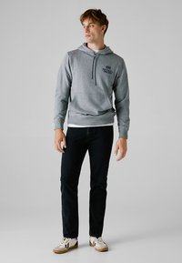 Sweatshirt cinzenta com capuz, cordão ajustável, bolso frontal e logótipo impresso. Combinada com jeans pretos e ténis brancos. Textura suave por todo o tecido.