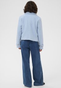 Personne portant une veste texturée bleu clair et un pantalon large en jean bleu, se tenant de dos face à l'appareil photo sur un fond uni.