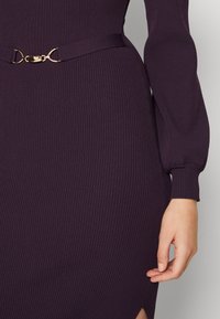 Gros plan sur une personne portant une robe côtelée violet foncé avec une ceinture à boucle dorée et des manches longues bouffantes, montrant la main et la zone de la taille.
