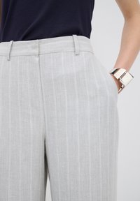 Lichtgrijze, gestreepte broek met een tailored fit, zijzakken en een gladde textuur. Gecombineerd met een zilveren manchetarmband.