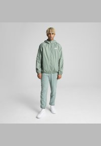 Lys grønn vindjakke med hette, elastiske mansjetter og hvite detaljer, paret med matchende joggebukser i en lignende nyanse og hvite sneakers.